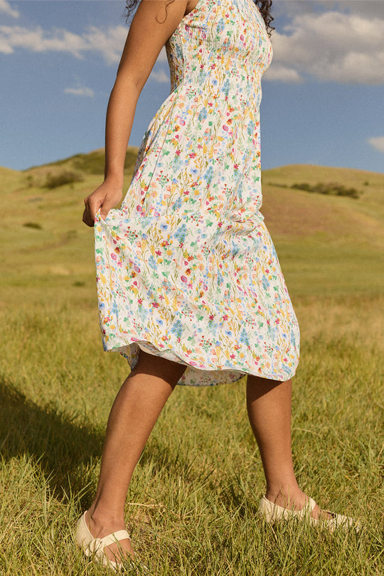 A person wearing the Ana Dress in Floral Fields by Ivy City Co, a colorful floral dress featuring a smocked bodice and a maternity-friendly design, walks on green grass in a sunny open field with hills and a blue sky in the background. This dress combines style and comfort, making it an ideal choice for expecting mothers.