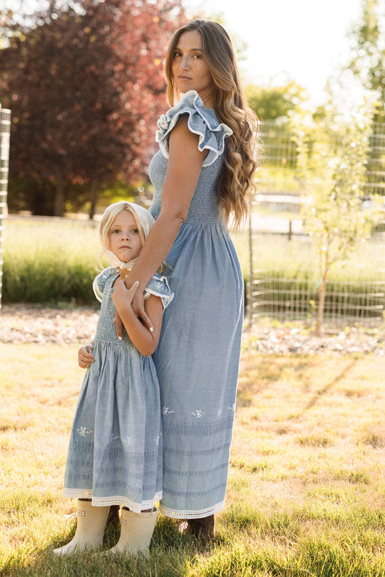 A woman and a young girl, both dressed in matching Ivy City Co Hattie Ankle Length Dresses in Embroidered Chambray featuring smocked bodices, stand together in a sunlit grassy yard. The woman holds the girl’s hand as they look toward the camera in these beautiful, maternity-friendly outfits.