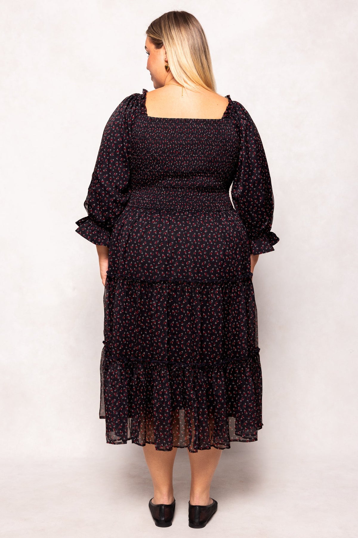 A woman with long blonde hair is standing with her back to the camera, wearing the Madeline Dress in Black Ditsy Floral by Ivy City Co—a black ditsy floral dress featuring long sleeves and an ankle-length hem, paired with black flats. This dress is both maternity-friendly and perfect for nursing mothers, offering comfort without sacrificing style.