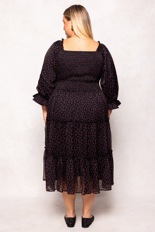 A woman with long blonde hair is standing with her back to the camera, wearing the Madeline Dress in Black Ditsy Floral by Ivy City Co—a black ditsy floral dress featuring long sleeves and an ankle-length hem, paired with black flats. This dress is both maternity-friendly and perfect for nursing mothers, offering comfort without sacrificing style.