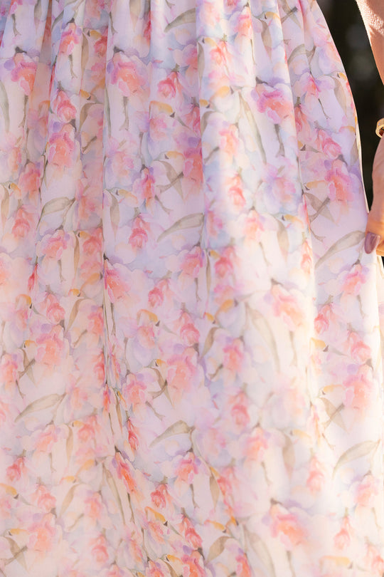Close-up of a flowing chiffon Kyla Dress in Watercolor Orchids by Ivy City Co, featuring a soft, pastel floral pattern in pink, purple, and green tones. A manicured hand adorned with a bracelet gently holds the edge of the nursing-friendly dress, highlighting its delicate elegance.