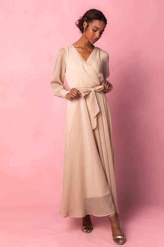 A woman wearing the Ivy City Co Andie Dress in Champagne—a long, light beige maxi dress with a tied waist and long sleeves—stands against a pink background, looking down as she adjusts the dress with one hand, paired with open-toe heels.