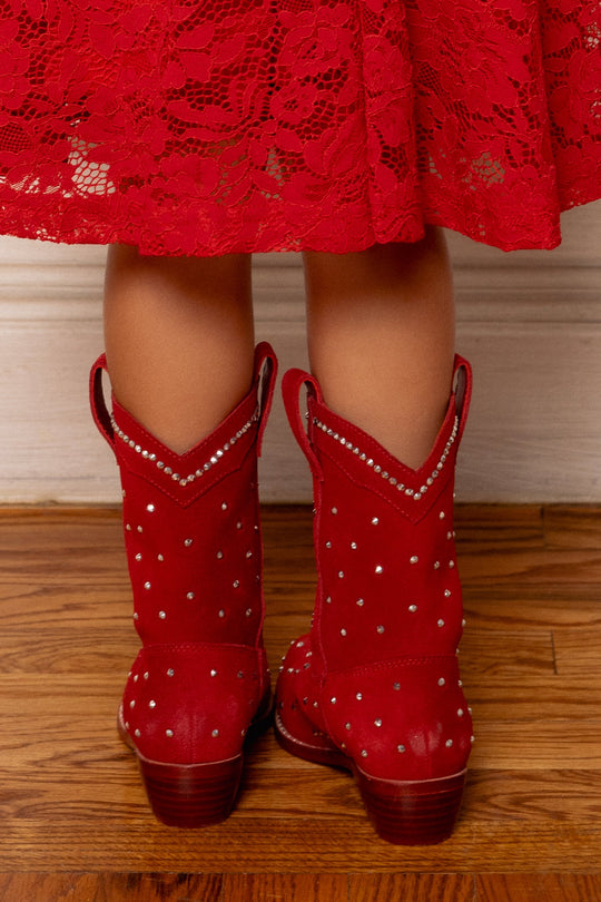 A child wearing a red lace dress and the Ivy City Co Mini Channing Bootie in Red, adorned with silver studs, stands on a wooden floor facing away from the camera.