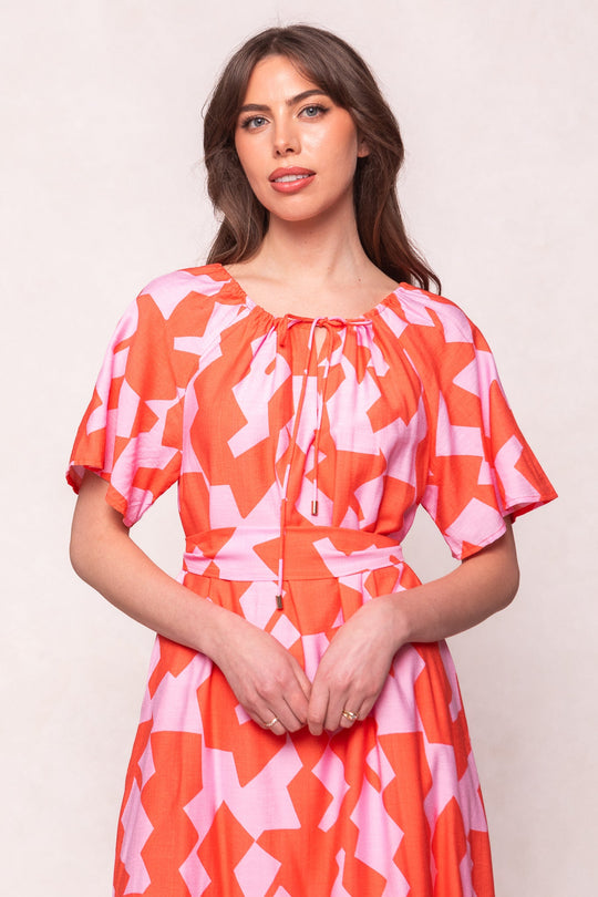 A woman stands against a light background, looking forward with hands clasped, wearing the Ivy City Co Haven Dress in Geometric—a red and pink geometric-patterned dress featuring short sleeves and a drawstring neckline.