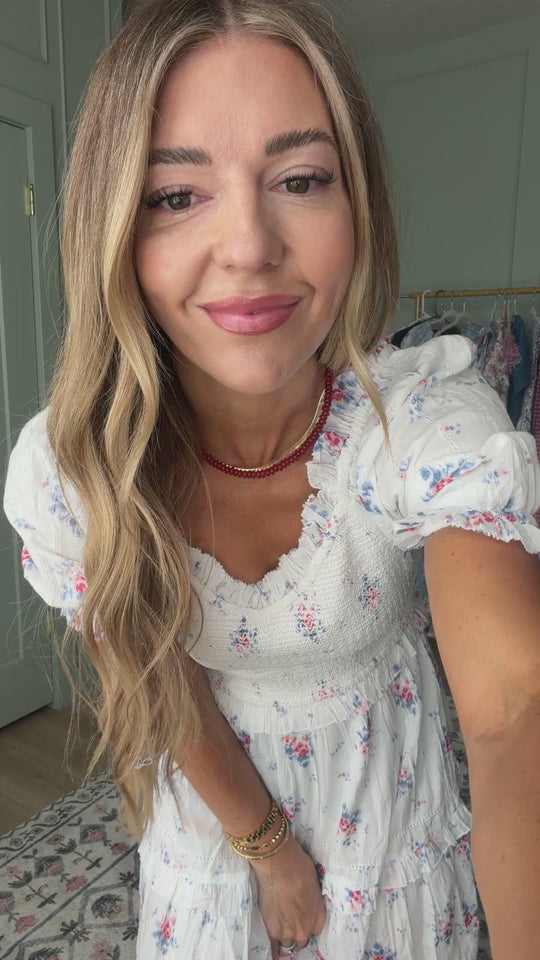 A woman with long, wavy blonde hair smiles at the camera, wearing the Emily Dress in Heritage Soft Ditsy Floral by Ivy City Co and a red beaded necklace, standing in a softly lit room.