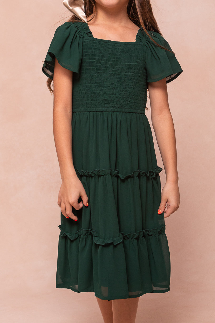 A girl wearing an Ivy City Co Mini Ellie Dress in Emerald Chiffon featuring an emerald smocked bodice, flutter sleeves, and a tiered, ruffled skirt.