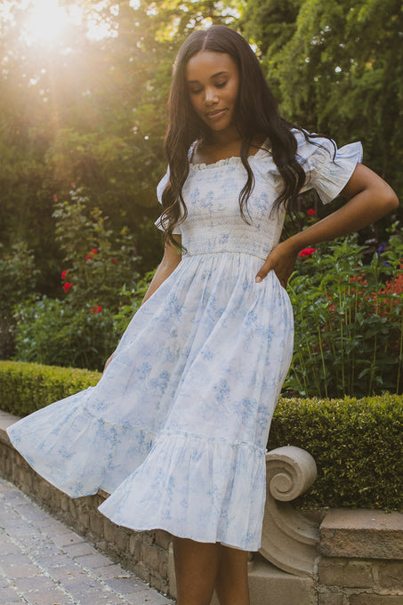 A woman in a light blue floral Valencia Dress by Ivy City Co stands near a garden, sunlight filtering through the trees behind her. She gently smiles, looking down while holding her cotton dress as she walks along a stone path.