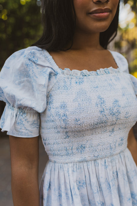 A woman wearing the Ivy City Co Valencia Dress, a light blue and white cotton maternity-friendly dress with puff sleeves, stands outdoors. The cropped image shows her from shoulders to waist with a softly blurred sunny background.
