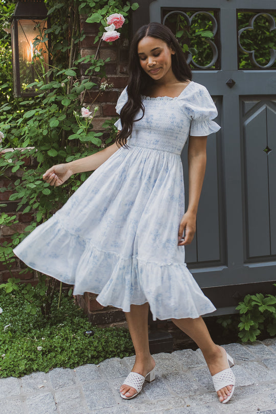 A woman in Ivy City Co’s light blue Valencia Dress, a short-sleeve, ruffled cotton style that’s maternity friendly, pairs it with white sandals as she stands smiling near a gray gate and blooming roses, holding her skirt.