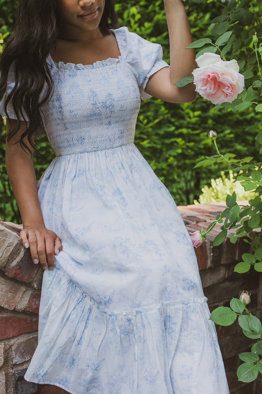 A woman in the Ivy City Co Valencia Dress, a light blue short-sleeved cotton style thats also maternity friendly, sits on a brick wall surrounded by green foliage and pink roses, gently touching a rose with one hand. Her face is partially out of frame.