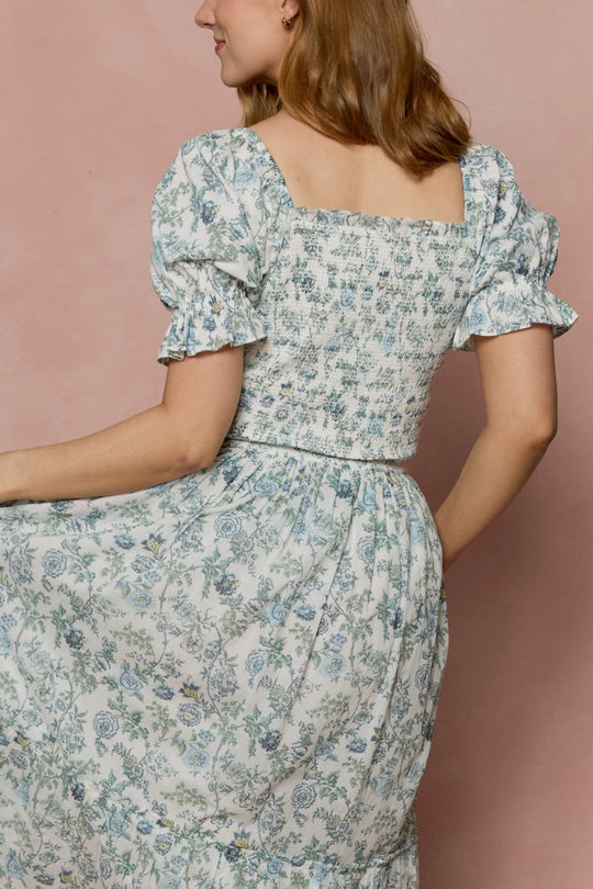 A woman, facing away and slightly to the side against a pink background, wears the Ivy City Co Valencia Dress in Ivory Climbing Vine Bloom—a floral cotton midi dress with puffed sleeves and a smocked back. This maternity-friendly style blends comfort and elegance, making it perfect for any occasion.