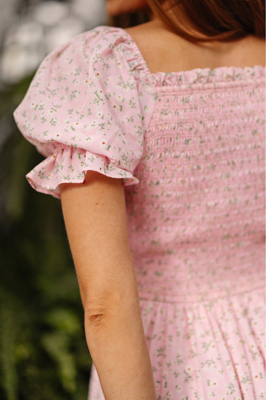 A person is shown from the back wearing Ivy City Co’s Valencia Dress in Pink Ditsy Floral, a maternity-friendly dress with ruffled short sleeves and a shirred bodice, set against softly blurred greenery.