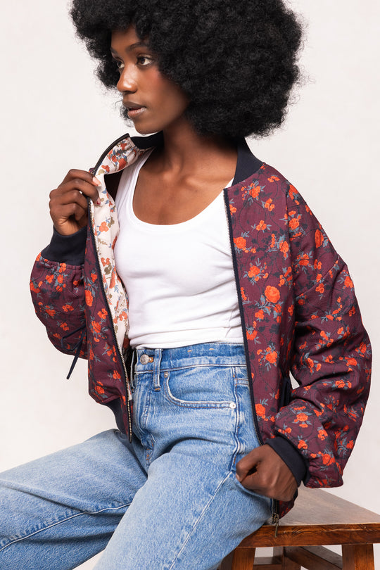Woman wearing a floral bomber jacket and jeans sitting on a wooden bench.