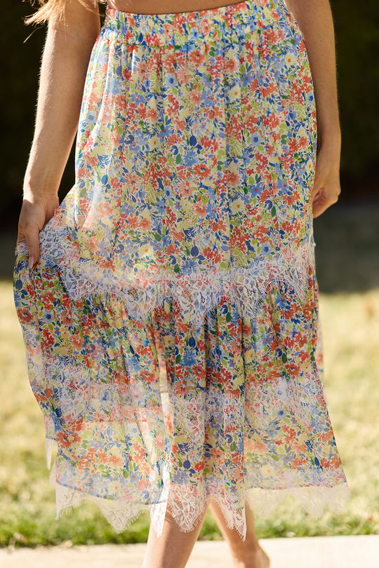 A person outdoors in sunlight holds out the Verona Skirt in Vibrant Yarrow by Ivy City Co, showcasing its colorful maxi silhouette and white lace trim against a backdrop of green grass and blurred foliage.