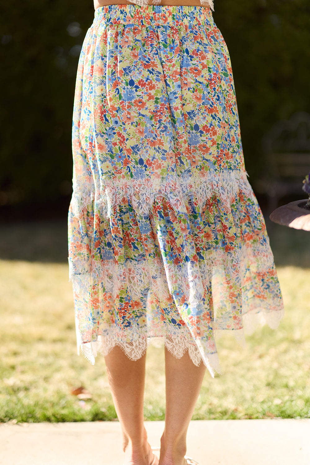 A person stands in a garden, wearing the Ivy City Co Verona Skirt in Vibrant Yarrow—a floral maxi style with white lace trim at the hem. Only their skirt and legs appear, set against sunlit greenery that highlights its vivid colors.
