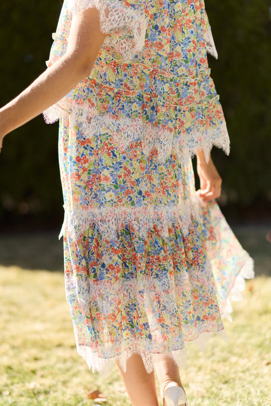 A person strolls on grass in the Ivy City Co Verona Skirt in Vibrant Yarrow, its colorful floral print and flowing silhouette glowing in sunlight, with greenery blurred behind, highlighting the skirt’s elegance.