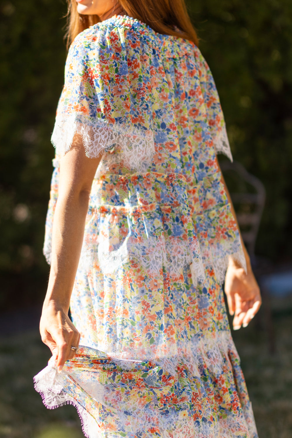 A woman wears the Ivy City Co Verona Top in Vibrant Yarrow, featuring lace trim and puff sleeves, outdoors. Sunlight highlights her brown hair as she gently holds the hem, with blurred greenery in the background.