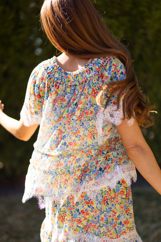 A woman with long brown hair stands outdoors, wearing Ivy City Co's Verona Top in Vibrant Yarrow with lace trim and puff sleeves, facing away with one arm raised against a blurred green backdrop.