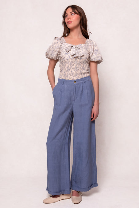 A woman stands against a light background in a puff-sleeve top with a bow and the Veronica Pants in Blue by Ivy City Co, featuring a high waist and wide-leg linen-blend design. With hands in her pockets, she embraces the relaxed summer style.