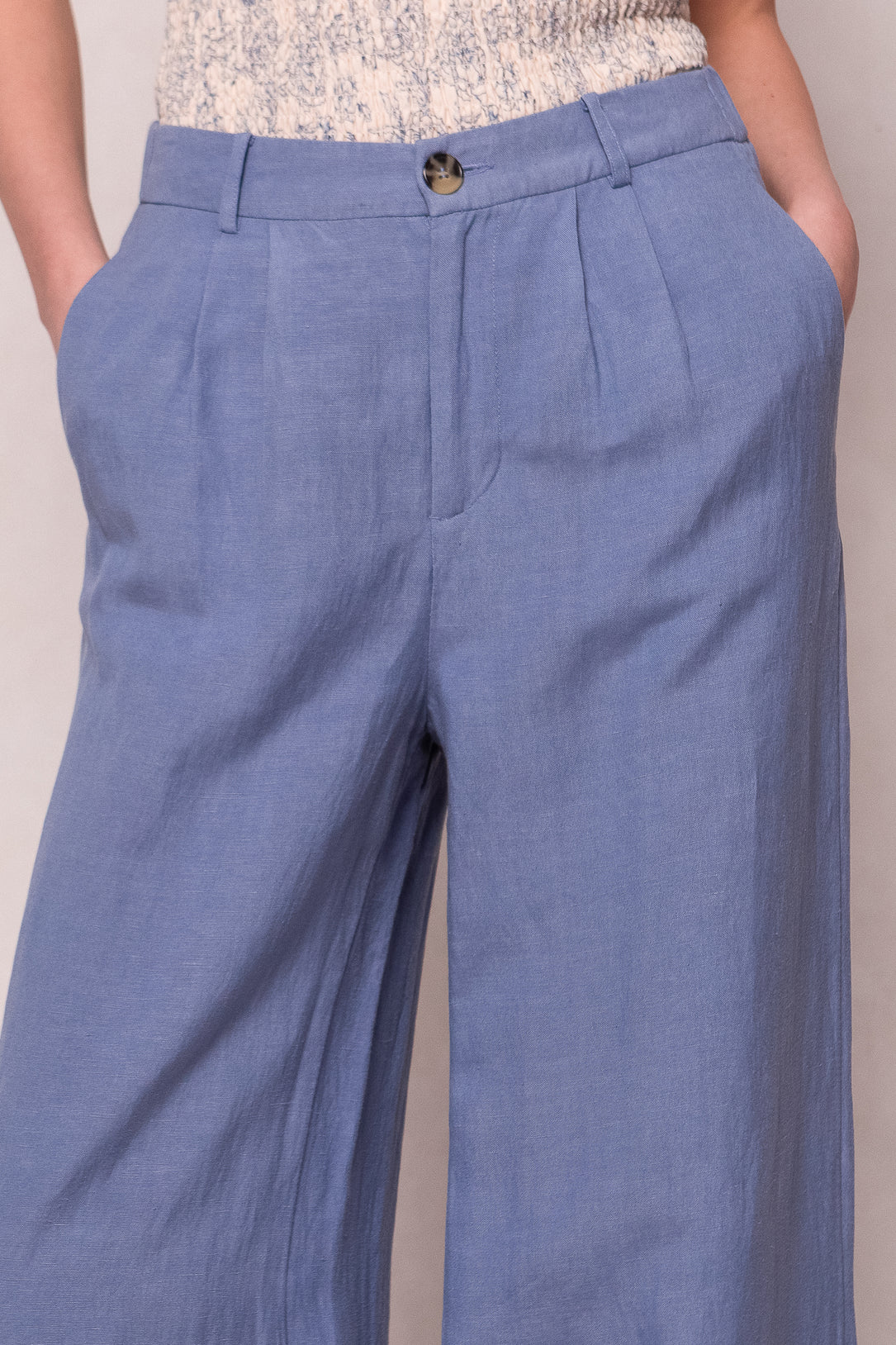 A person wears the Veronica Pants in Blue by Ivy City Co—high-waisted, light blue wide-leg linen-blend pants with pleats, side pockets, button closure, and hands in pockets. The top of a light patterned blouse is also seen.
