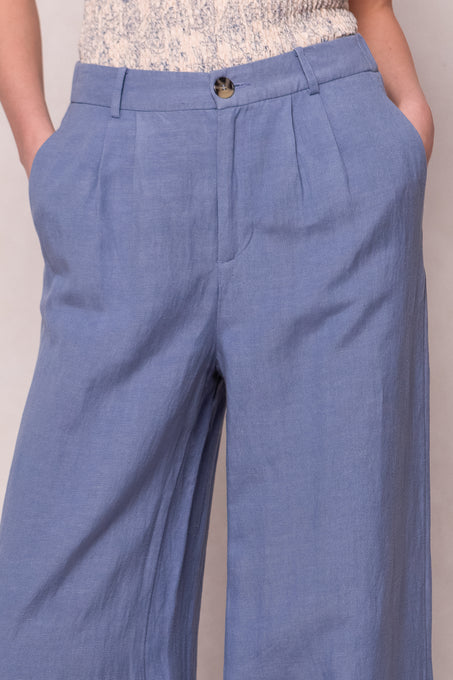 A person wears the Veronica Pants in Blue by Ivy City Co—high-waisted, light blue wide-leg linen-blend pants with pleats, side pockets, button closure, and hands in pockets. The top of a light patterned blouse is also seen.