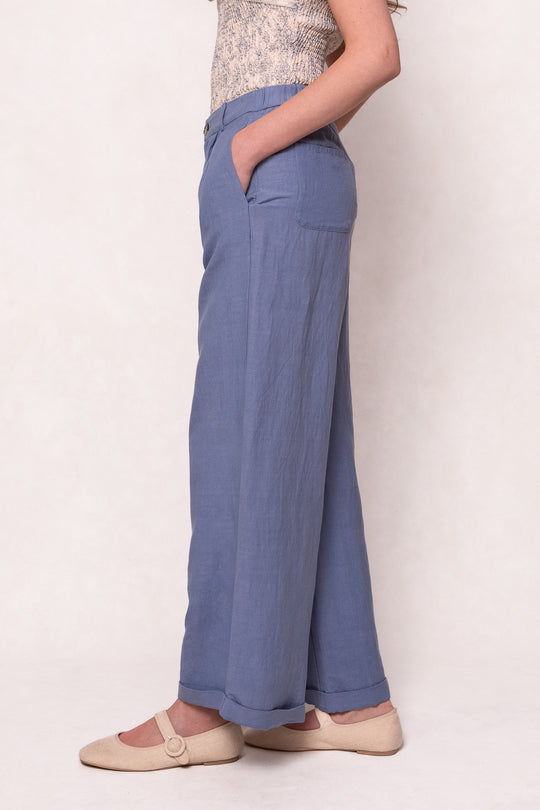 A person stands in profile with one hand in their pocket, wearing Ivy City Cos Veronica Pants in Blue—a relaxed-fit linen-blend—paired with a patterned top and beige strappy flats against a plain light background.