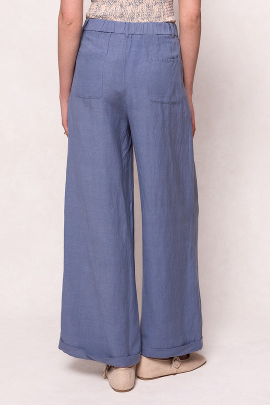 Someone is seen from behind wearing Ivy City Co’s Veronica Pants in Blue—a relaxed-fit, linen-blend style with back pockets—paired with beige shoes, all set against a light background.