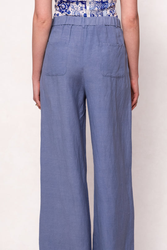 A person is seen from the back wearing Ivy City Co’s Veronica Pants in Blue—relaxed fit, high-waisted, linen-blend trousers with an elastic waistband and two back pockets—paired with a patterned top.