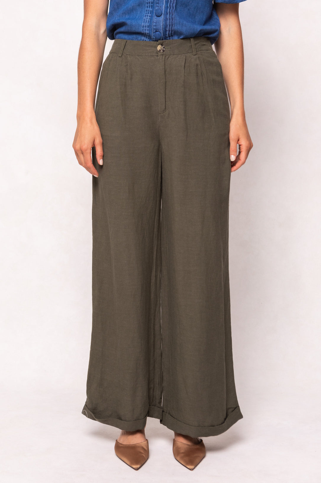 A person is shown from shoulders to feet against a light background, wearing Ivy City Co’s Veronica Pants in Green—high-waisted and wide-leg—paired with a tucked-in blue denim shirt and brown pointed-toe shoes.