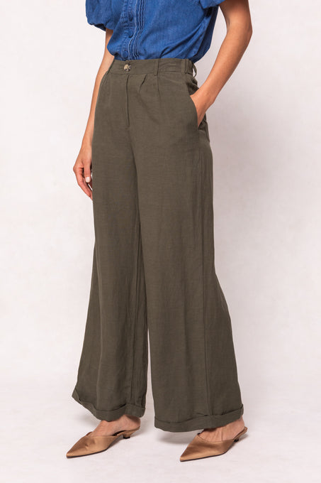 A person models the Ivy City Co Veronica Pants in Green—wide-leg, linen-blend with an elastic waistband—paired with a blue short-sleeve top and brown pointed-toe flats, posing against a plain light background.