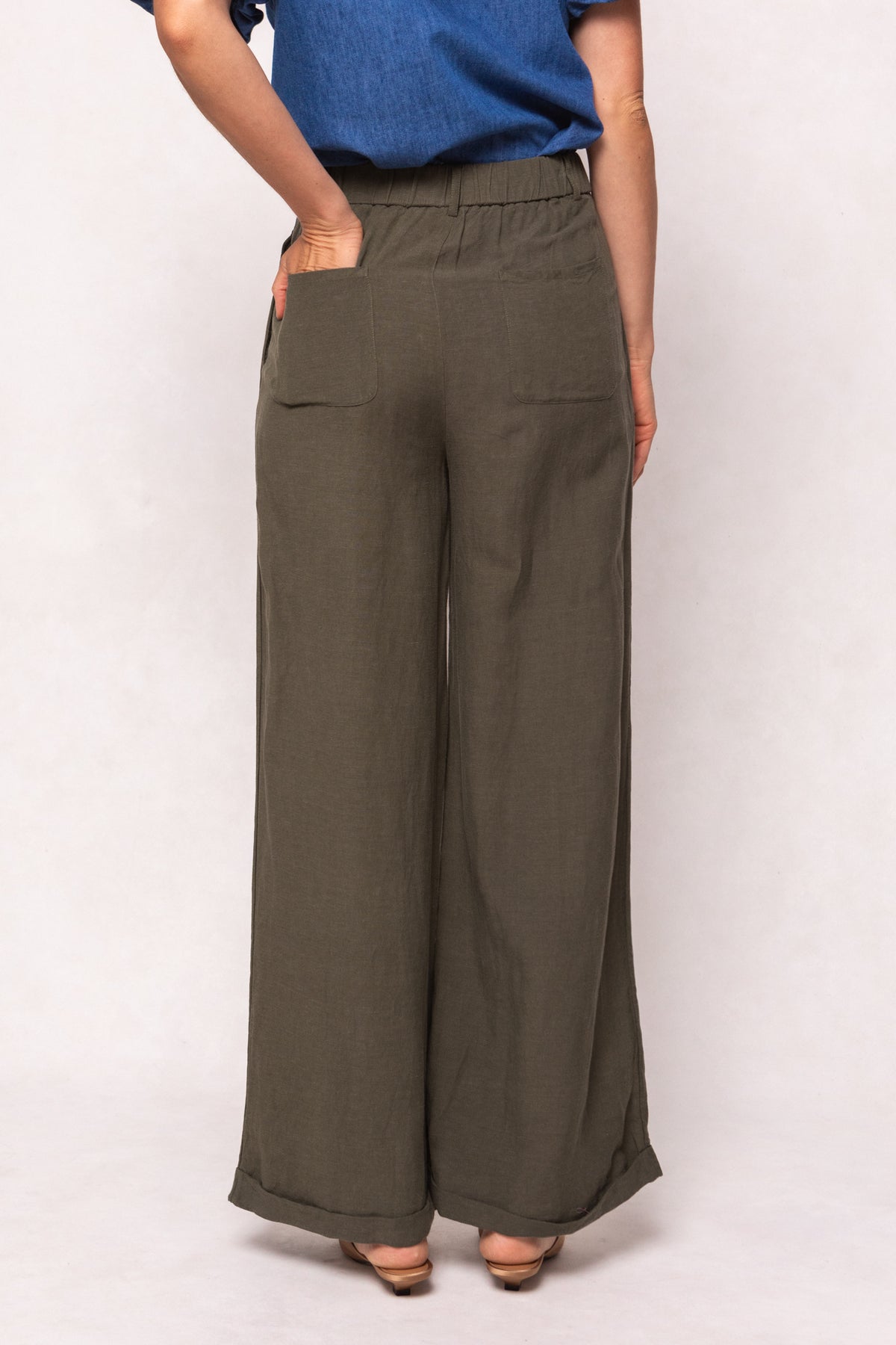 A person is seen from behind wearing Ivy City Cos Veronica Pants in Green—loose-fitting, wide-leg linen-blend trousers with back pockets and an elastic waistband—paired with a blue short-sleeved top against a light background.