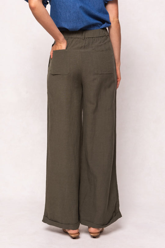 A person is seen from behind wearing Ivy City Cos Veronica Pants in Green—loose-fitting, wide-leg linen-blend trousers with back pockets and an elastic waistband—paired with a blue short-sleeved top against a light background.