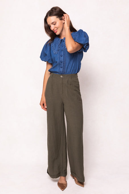 A woman stands against a light background, smiling gently as she touches her hair. She wears Ivy City Cos Veronica Pants in Green—high-waisted, wide-leg trousers with an elastic waistband—paired with a blue ruffled blouse and tan pointed-toe flats.