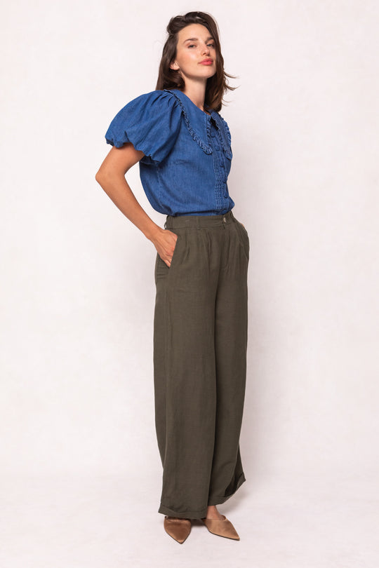 A woman stands sideways against a light background, wearing a blue puff-sleeve blouse, Ivy City Co Veronica Pants in Green (high-waisted linen-blend wide-leg trousers), and brown pointed flats.