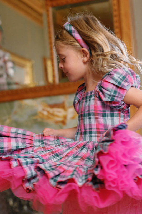 A young girl wearing Ivy City Co’s Hair Bows in Vibrant Plaid twirls joyfully in a pink and green plaid dress with puffed sleeves, her skirt and tulle layers swirling outward, as she appears delighted in a room adorned with a large ornate mirror.