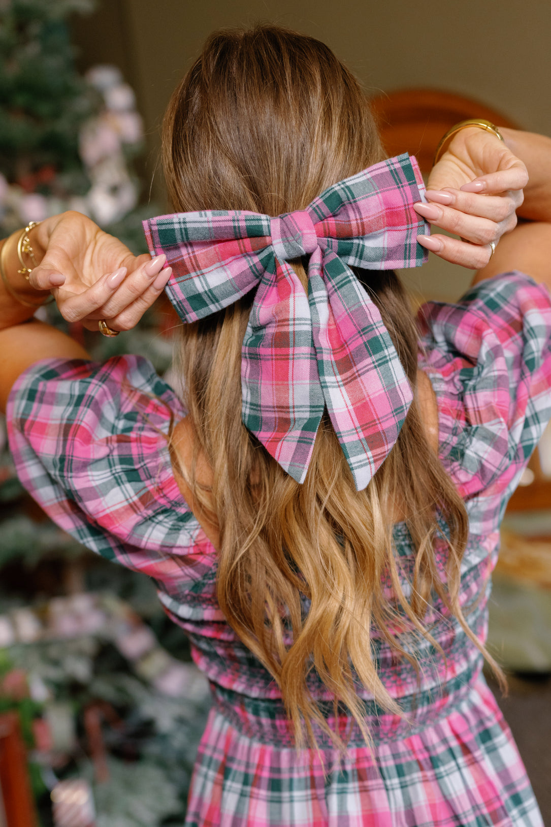 A woman with long wavy hair wears an Ivy City Co Hair Bow in Vibrant Plaid, perfectly matching her dress as she holds the festive holiday accessory with both hands. A decorated Christmas tree is softly blurred in the background.