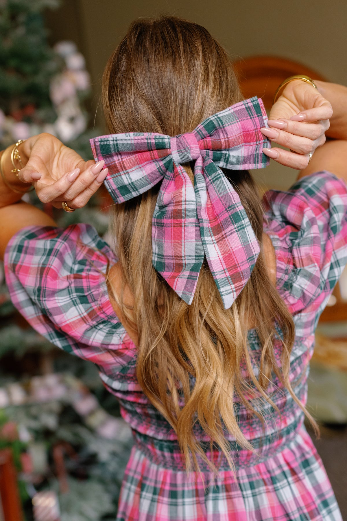 A woman with long wavy hair wears an Ivy City Co Hair Bow in Vibrant Plaid, perfectly matching her dress as she holds the festive holiday accessory with both hands. A decorated Christmas tree is softly blurred in the background.