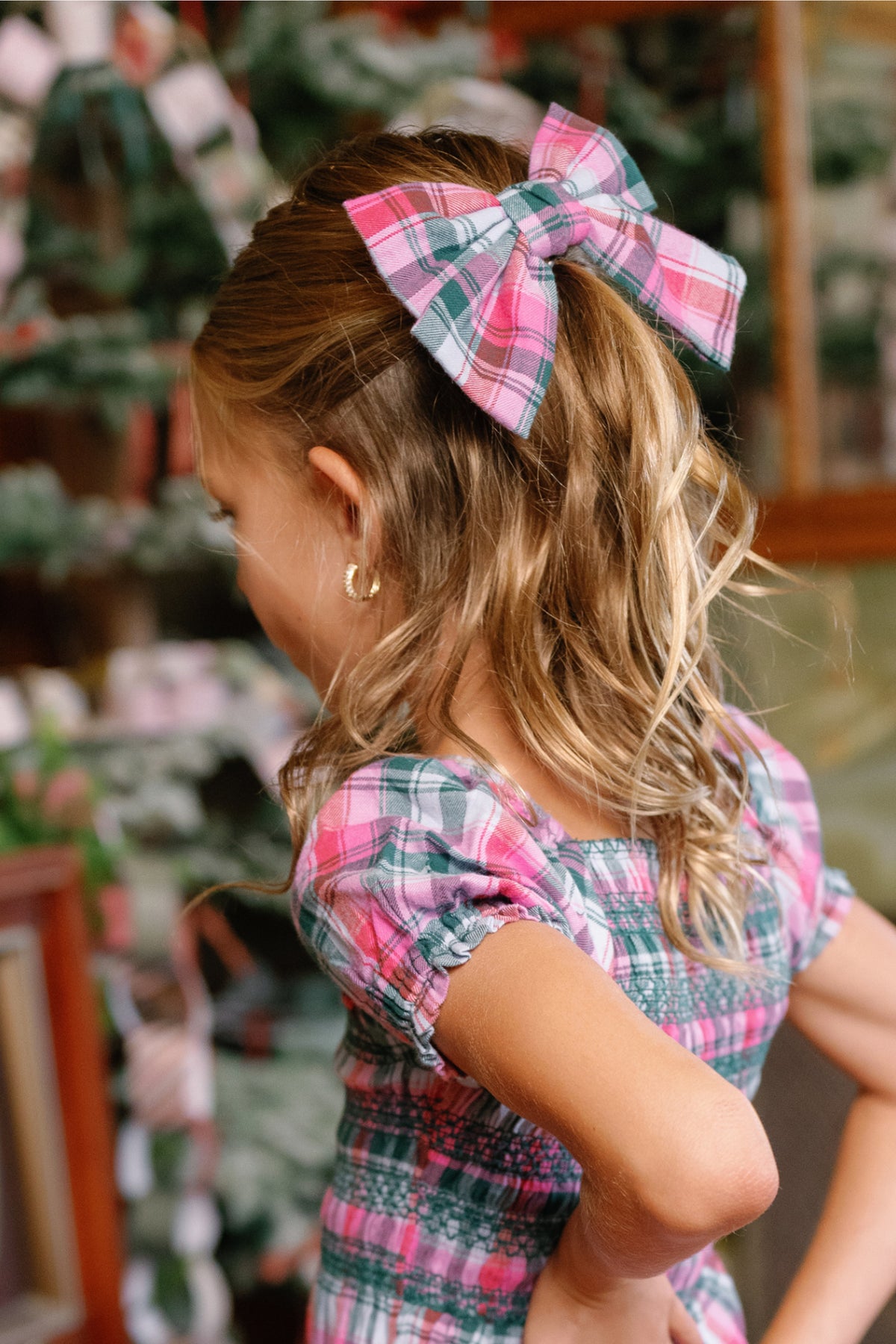 A young girl with wavy blond hair wears an Ivy City Co Hair Bow in Vibrant Plaid—a delightful holiday accessory—paired with a matching plaid dress. Standing with her hands on her hips and facing away from the camera, she is set against a backdrop of festive holiday paper chains.