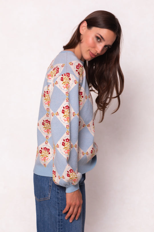 A woman with long brown hair wears the Ivy City Co Vivienne Sweater in Mosaic Bloom, a light blue maternity-friendly sweater with a floral diamond pattern, paired with jeans, standing sideways and looking at the camera against a light background.