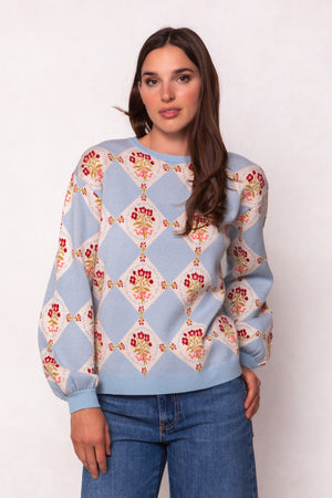 A woman with long brown hair wears Ivy City Cos Vivienne Sweater in Mosaic Bloom, a light blue maternity-friendly sweater with diamond and floral patterns, paired with blue jeans against a light neutral background.