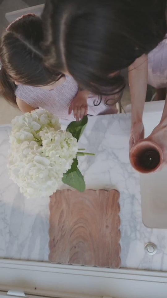 A woman wearing the Ivy City Co Karla Dress in Pink Striped Gingham holds a cup as she stands beside a girl at a marble counter adorned with a vase of white hydrangeas and a wooden cutting board.