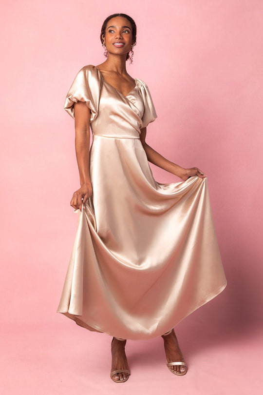 A woman poses in the Ivy City Co Tessie Dress in Champagne—a shiny satin wedding guest dress with puff sleeves—holding out the skirt. She stands on a pink background, smiling and wearing open-toe heels.