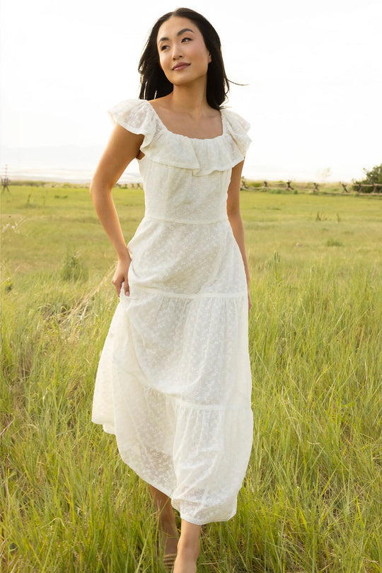 A woman wearing the Alaina Dress in Ivory by Ivy City Co, featuring a delicate smocked bodice and ruffled, maxi-length silhouette, stands content and relaxed in a grassy field on a sunny day.