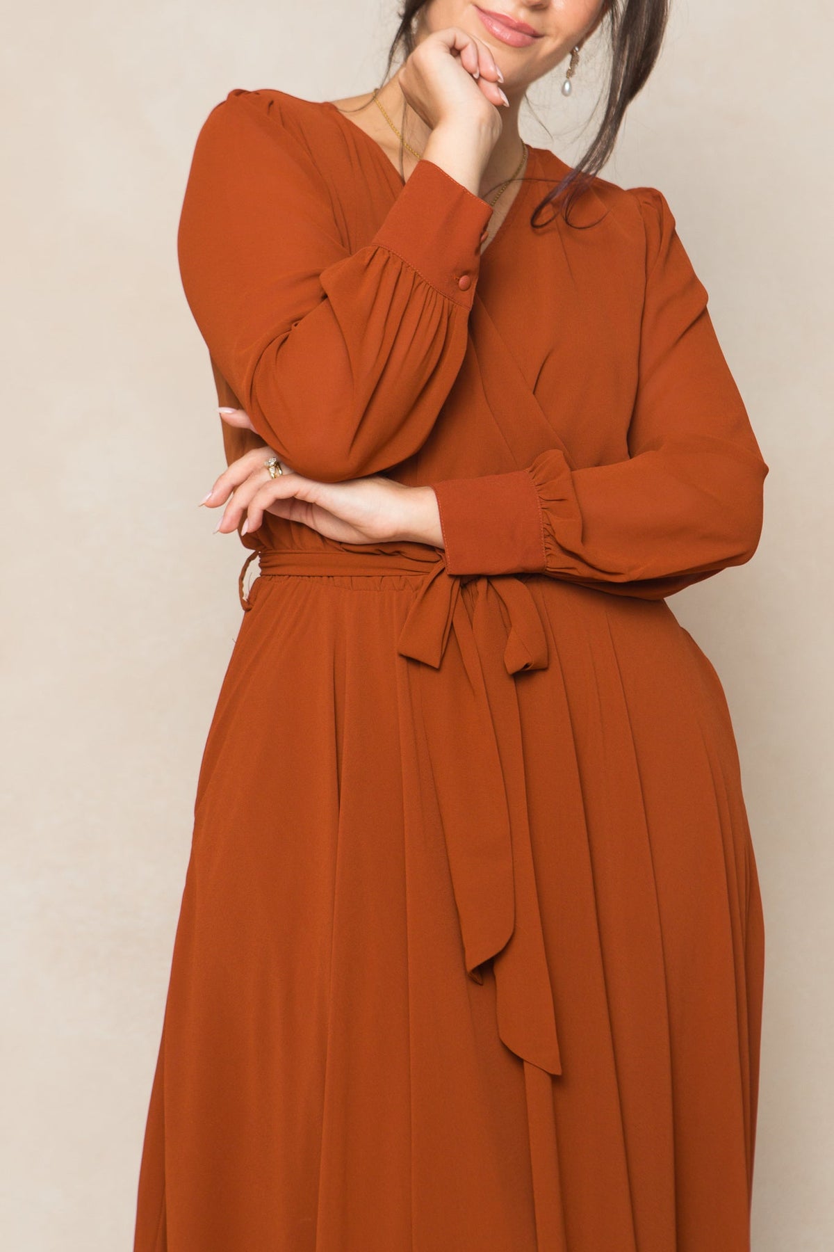 A woman wearing the Andie Dress in Amber Chiffon by Ivy City Co— a long-sleeved women’s maxi dress with a waist tie —stands with one hand resting on her chin and the other arm crossed. Her face is partially visible, set against a neutral beige background.