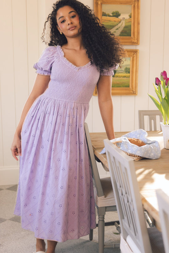Wearing the Emmi Dress in Lilac by Ivy City Co—a maternity friendly eyelet cotton style—a woman with curly hair stands beside a dining table adorned with flowers, a basket, and a blue cloth. Behind her, framed landscape paintings hang on the wall.
