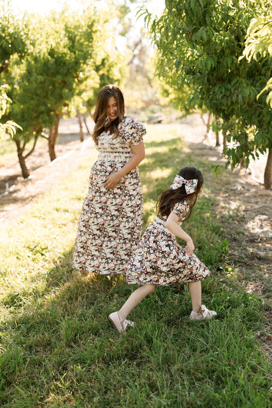 A woman and a young girl wearing matching Mini Sunday Best Dresses in Midnight Garden by Ivy City Co, with puff sleeves and floral prints, walk and play together in a sunlit orchard surrounded by green trees and grass.