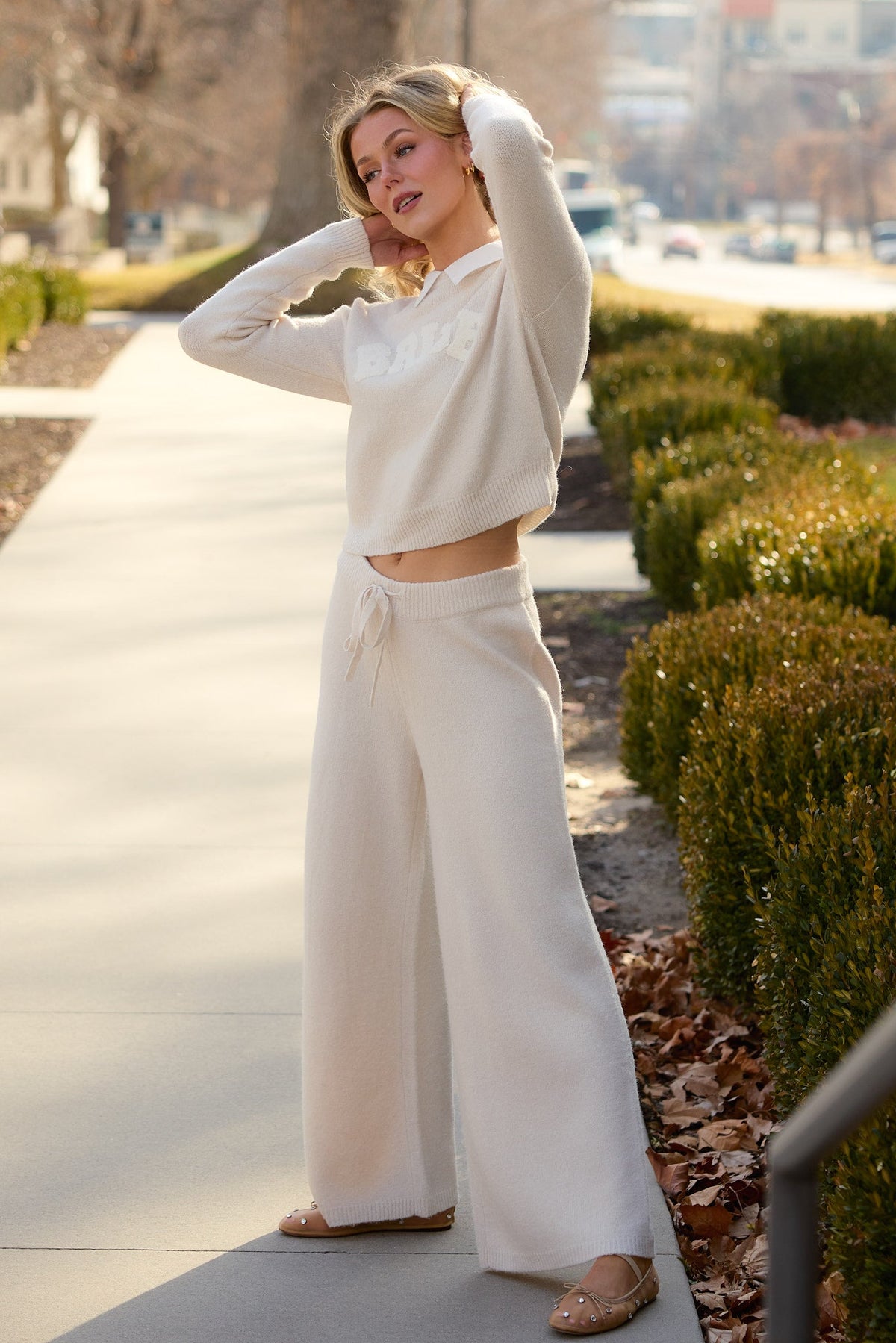 A woman stands on a sidewalk surrounded by greenery on a sunny day, wearing the Ivy City Co Bride Sweater in Oatmeal with wide-leg pants and nude flats. She has her hands behind her head.