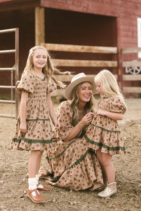 A woman kneels between two young girls outdoors, all wearing matching Ivy City Co Mini Roselyn Dresses in Desert Blossom, smiling together in front of a wooden fence and barn.