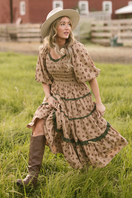 A woman wearing the Ivy City Co Roselyn Dress in Desert Blossom stands in a grassy field near a wooden fence and red barn, styled with brown knee-high boots and a wide-brimmed hat.