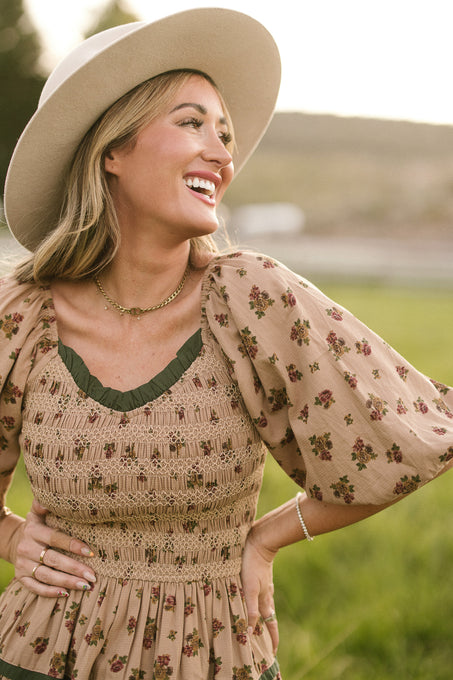 A smiling woman stands in a grassy field with sunlight on her face, wearing the Ivy City Co Roselyn Dress in Desert Blossom and a beige hat, her hand on her hip.